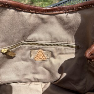 May include: A brown leather tote bag with a beige fabric interior. The bag features a zippered pocket with a gold-coloured zipper and a leather patch with the letters "BA" inside. The top edge of the bag is trimmed with dark brown leather.