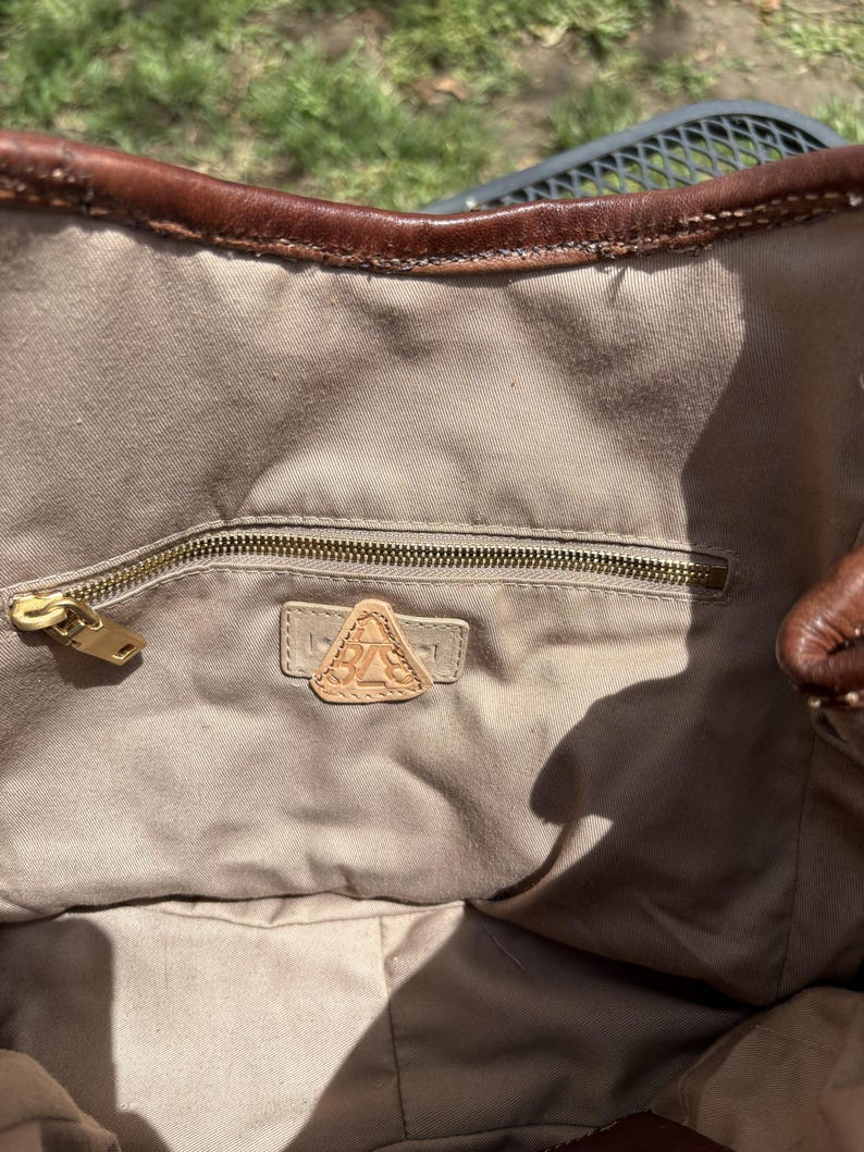 May include: A brown leather tote bag with a beige fabric interior. The bag features a zippered pocket with a gold-coloured zipper and a leather patch with the letters "BA" inside. The top edge of the bag is trimmed with dark brown leather.