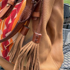 May include: A tan leather handbag with brown leather straps and decorative tassels. The bag features a patterned fabric panel with red, orange, and yellow geometric shapes. The bag is sitting on a metal table.