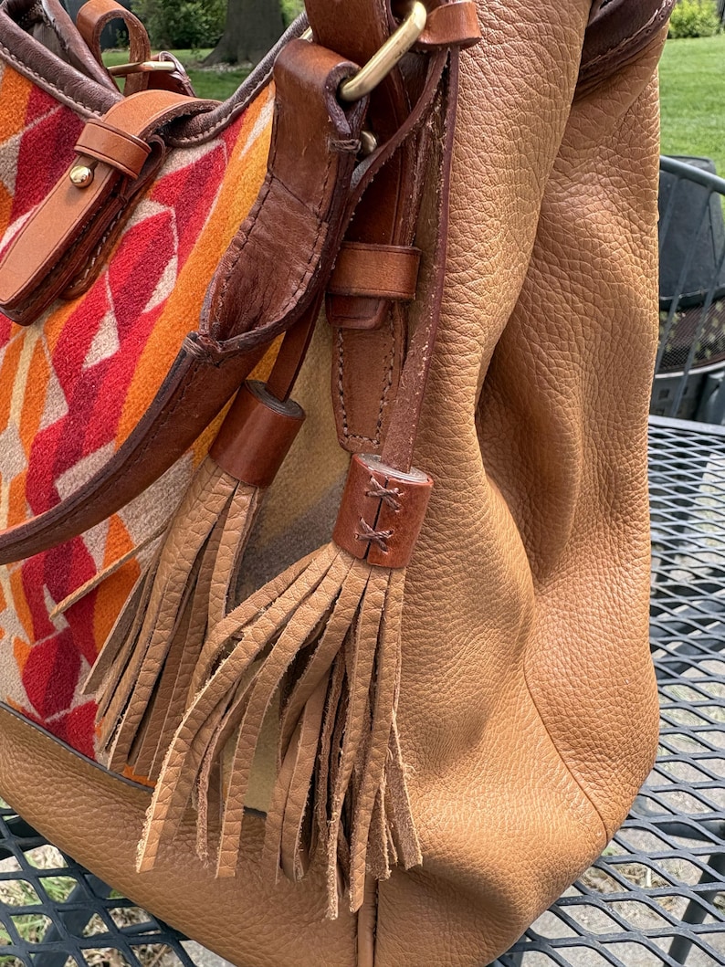 May include: A tan leather handbag with brown leather straps and decorative tassels. The bag features a patterned fabric panel with red, orange, and yellow geometric shapes. The bag is sitting on a metal table.
