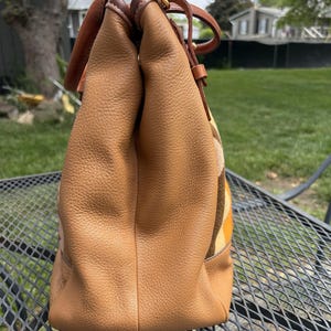 May include: A tan leather tote bag with brown leather handles and accents. The bag has a textured surface and a patterned fabric panel in shades of yellow, orange, and brown. The bag is sitting on a metal table.