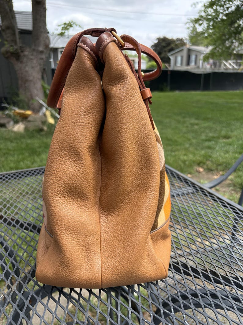 May include: A tan leather tote bag with brown leather handles and accents. The bag has a textured surface and a patterned fabric panel in shades of yellow, orange, and brown. The bag is sitting on a metal table.