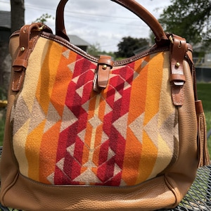 May include: A brown leather handbag with a geometric patterned front in shades of red, orange, and tan. The bag has a top handle, leather accents, and a tassel.