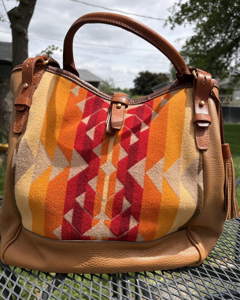 May include: A brown leather handbag with a geometric patterned front in shades of red, orange, and tan. The bag has a top handle, leather accents, and a tassel.