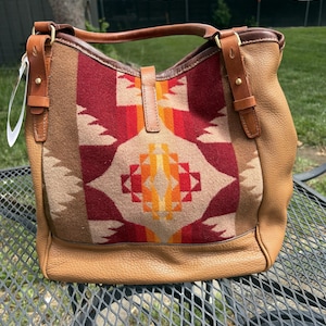 May include: A tan leather handbag with a patterned wool panel. The panel features a geometric design in shades of red, orange, and beige. The bag has a top handle and adjustable shoulder straps, with brass-coloured hardware.