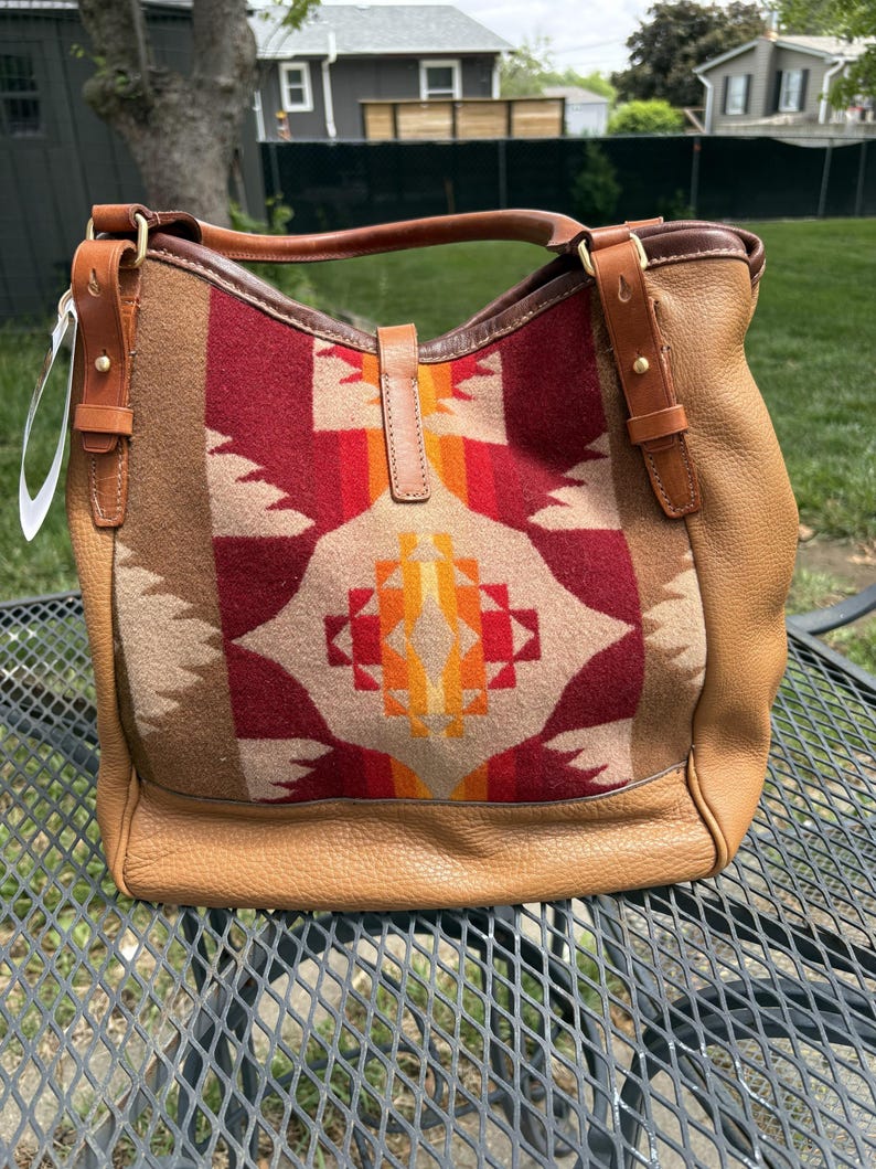 May include: A tan leather handbag with a patterned wool panel. The panel features a geometric design in shades of red, orange, and beige. The bag has a top handle and adjustable shoulder straps, with brass-coloured hardware.