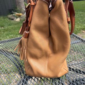 May include: A tan leather handbag with a textured finish. The bag has brown leather details, a shoulder strap, and a decorative tassel. The bag is on a metal table.