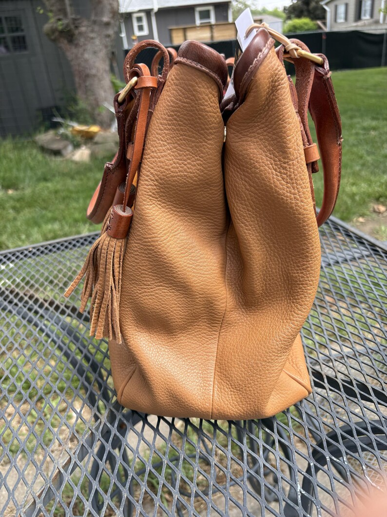 May include: A tan leather handbag with a textured finish. The bag has brown leather details, a shoulder strap, and a decorative tassel. The bag is on a metal table.