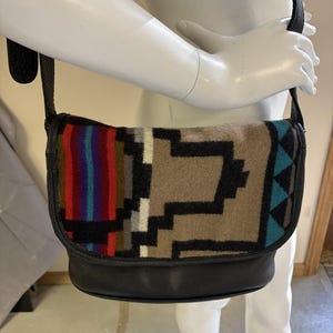 May include: A black leather crossbody bag with a flap closure. The bag features a colorful woven fabric panel with geometric patterns in red, blue, brown, and black. The bag has a black leather strap.