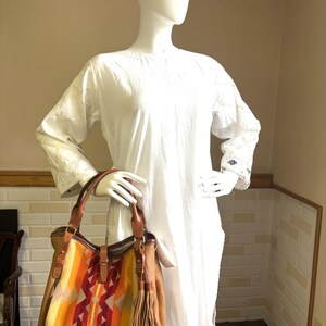 May include: A tan leather tote bag with a colourful woven panel featuring orange, yellow, and red geometric patterns. The bag has brown leather handles and a tassel. The bag is displayed on a white mannequin.