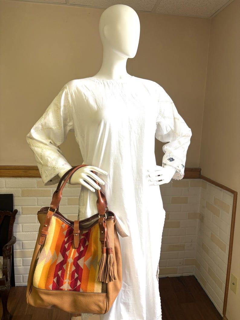 May include: A tan leather tote bag with a colourful woven panel featuring orange, yellow, and red geometric patterns. The bag has brown leather handles and a tassel. The bag is displayed on a white mannequin.