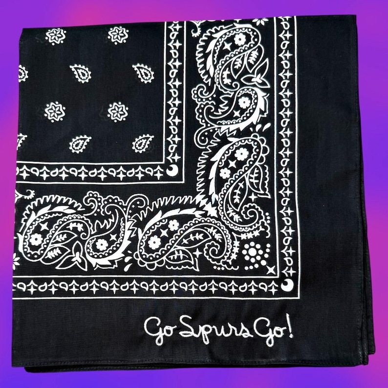 May include: A black bandana featuring a white paisley pattern with floral and star motifs. The fabric is bordered by a decorative line of small crosses and teardrop shapes. The text Go Spurs Go! is printed in white script in the bottom corner.
