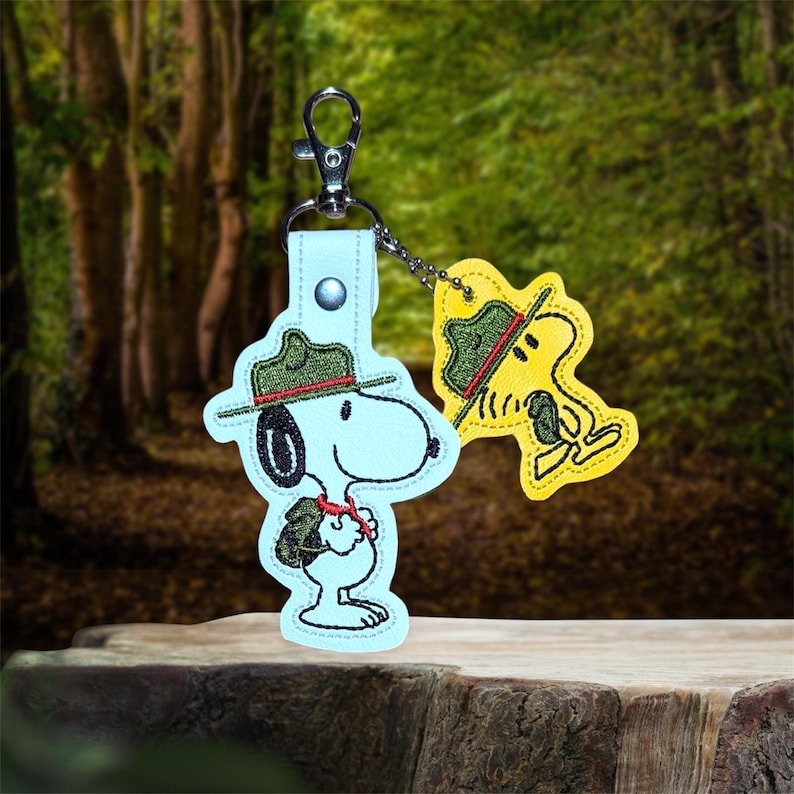 Puede incluir: Un llavero de Snoopy azul claro con un sombrero de guardabosques verde y rojo y un llavero de Woodstock amarillo. Los llaveros est&aacute;n sujetos a un cierre de metal plateado. El fondo es una escena de bosque borrosa.