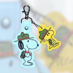 Puede incluir: Un llavero de Snoopy azul claro con un cierre plateado. El llavero presenta un dise&ntilde;o bordado de Snoopy con un sombrero verde y un pa&ntilde;uelo rojo. Un llavero de Woodstock amarillo m&aacute;s peque&ntilde;o est&aacute; adjunto.