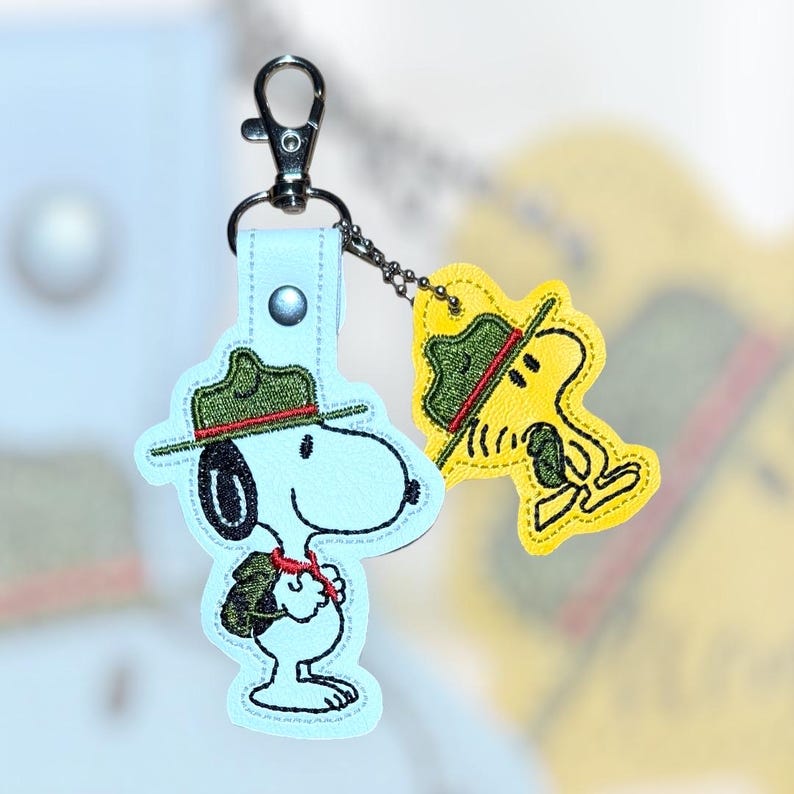 Puede incluir: Un llavero de Snoopy azul claro con un cierre plateado. El llavero presenta un dise&ntilde;o bordado de Snoopy con un sombrero verde y un pa&ntilde;uelo rojo. Un llavero de Woodstock amarillo m&aacute;s peque&ntilde;o est&aacute; adjunto.