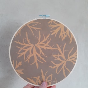 May include: A round embroidery hoop featuring a brown fabric with a repeating pattern of light orange botanical shapes. The wooden hoop is secured with a metal screw and held by a hand.