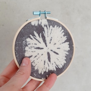 May include: A white and gray floral design on a round piece of fabric stretched in a wooden embroidery hoop.
