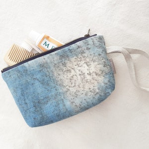 Puede incluir: Una bolsa de cremallera de mezclilla azul con un asa de tela blanca. La bolsa tiene un aspecto descolorido y desgastado y está llena de un tubo de pasta de dientes y un peine de madera.