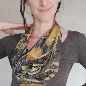Pode incluir: Um lenço de seda com um padrão de folhas em tons de castanho, dourado e verde. O lenço está drapeado ao redor do pescoço. O fundo é uma parede cinza-claro lisa.