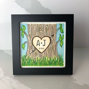 May include: A framed illustration of a tree with a heart carved into the trunk. The heart is light brown with the initials "A+J" and the date "2.4.21" inside. The tree is brown with green leaves and grass at the base.