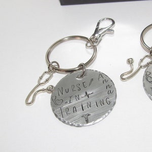 May include: Two silver keychains with a stethoscope charm. One keychain has a round tag that says "Nurse in Training" with a heart and a caduceus symbol. The other keychain has a round tag that says "Nurse in Training" with a heart, a caduceus symbol, and the name "Anna".