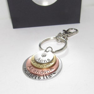 May include: A silver key chain with four stacked metal discs. The top disc is gold and says "Anna" with a heart symbol. The second disc is brass and says "Adams". The third disc is copper and says "Landon". The bottom disc is silver and says "Gabrielle Haas".