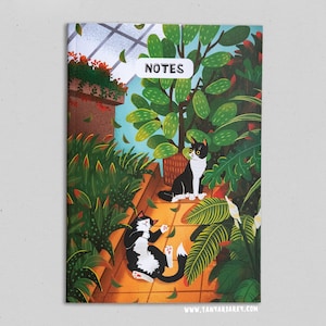 May include: A notebook cover featuring an illustration of two black and white cats in a lush green indoor garden setting. The text "NOTES" is printed in white on a black rectangle at the top of the cover. The bottom of the cover has the website "www.tanyajarikey.com" printed in white.