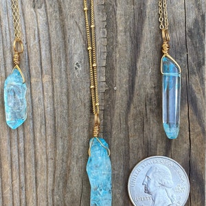 Puede incluir: Tres collares de oro con colgantes de cristal azul. Los colgantes est&aacute;n envueltos en alambre dorado y son de diferentes tama&ntilde;os. Los collares est&aacute;n sobre un fondo de madera con una moneda de un cuarto de d&oacute;lar estadounidense para la escala.