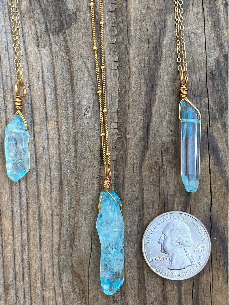 Puede incluir: Tres collares de oro con colgantes de cristal azul. Los colgantes est&aacute;n envueltos en alambre dorado y son de diferentes tama&ntilde;os. Los collares est&aacute;n sobre un fondo de madera con una moneda de un cuarto de d&oacute;lar estadounidense para la escala.