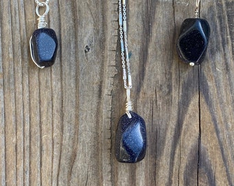 May include: Three silver necklaces with dark blue gemstone pendants. The pendants are different shapes and sizes. The necklaces are on a wooden background.