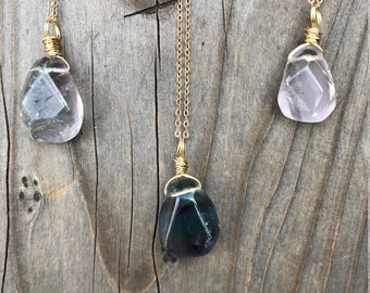 May include: Three gold necklaces with different colored crystal pendants. The pendants are wrapped in gold wire and are shaped like teardrops. The crystals are a light purple, a dark purple, and a light pink.