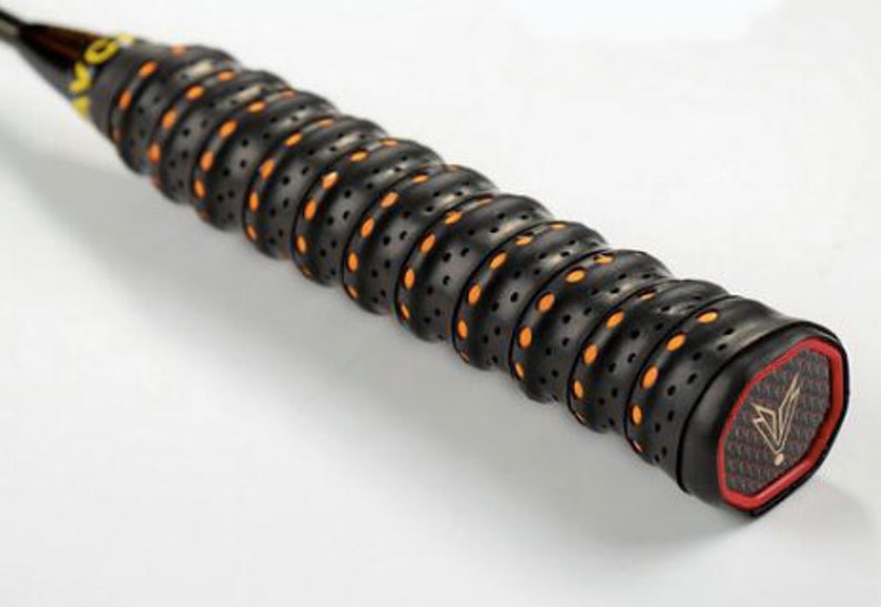May include: Close-up of a black badminton racket grip with orange and black patterned grip tape. The grip has a textured surface with small holes and a red and black end cap with a logo.