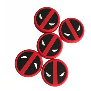 May include: Five red and black round stickers with a white Deadpool logo. The logo features a red slash across a black background with two white eyes.