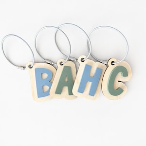 May include: Four wooden letter tags spelling B, A, H, and C, each attached to a flexible silver metal cable loop. The letters B and H are painted in a muted blue, while the letters A and C are painted in a soft sage green on light wood.