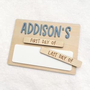 May include: A rectangular wooden sign featuring the blue 3D text ADDISON'S at the top. Below, two removable wooden strips are included, one labeled FIRST DAY OF and the other labeled LAST DAY OF, positioned over a white rectangular writing area.