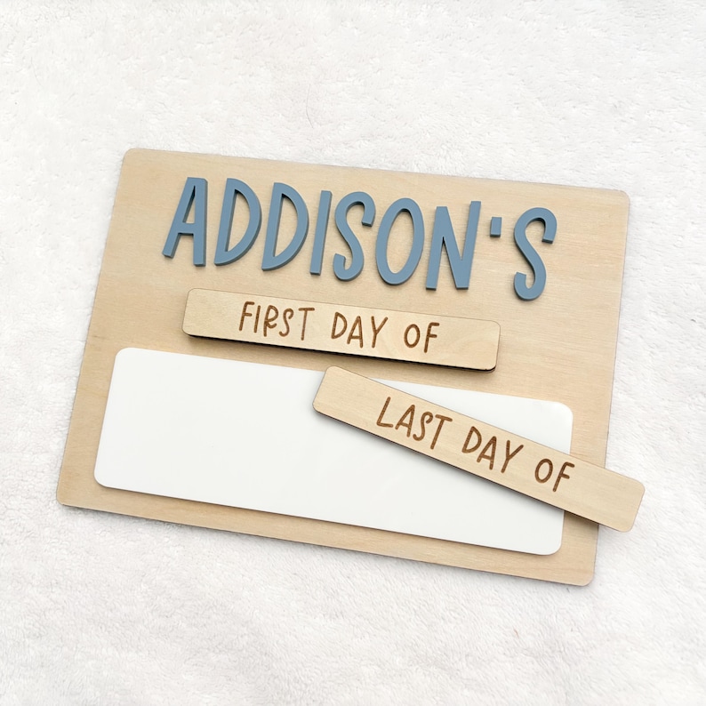 May include: A rectangular wooden sign featuring the blue 3D text ADDISON'S at the top. Below, two removable wooden strips are included, one labeled FIRST DAY OF and the other labeled LAST DAY OF, positioned over a white rectangular writing area.
