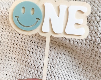 May include: A wooden cake topper with a light blue smiley face and the word "ONE" in white. The topper is on a wooden stick, perfect for a first birthday celebration. The background is a cream-colored knitted fabric.