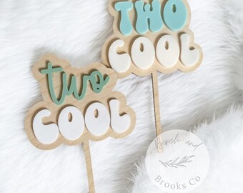 May include: Two wooden cake toppers with the words "two cool" in green and white letters. The toppers are on wooden sticks and are perfect for a birthday party.