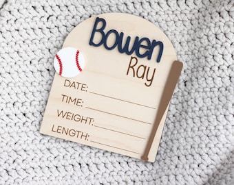 May include: A wooden baseball-themed birth announcement sign. The sign features the name "Bowen Ray" in navy blue and brown lettering, a baseball, and a baseball bat. It includes lines for date, time, weight, and length.