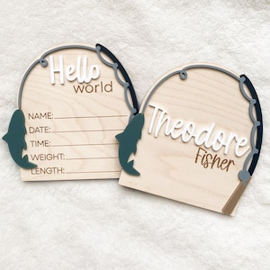 K&ouml;nnte beinhalten: Zwei Holzschilder mit Angelmotiv. Eines zeigt "Hello world" mit Zeilen zum Ausf&uuml;llen der Baby-Informationen. Das andere Schild zeigt "Theodore Fisher". Beide haben ein graues Angelruten-Design und einen gr&uuml;nen Fisch.