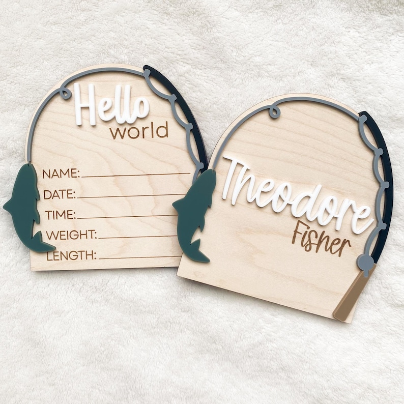 K&ouml;nnte beinhalten: Zwei Holzschilder mit Angelmotiv. Eines zeigt "Hello world" mit Zeilen zum Ausf&uuml;llen der Baby-Informationen. Das andere Schild zeigt "Theodore Fisher". Beide haben ein graues Angelruten-Design und einen gr&uuml;nen Fisch.