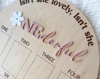 May include: A wooden round growth chart with the words "Isn't she lovely, Isn't she wonderful" written in brown. The chart has six squares with the numbers one through six written in black. A pink flower with the word "ONE" in pink letters is attached to the chart.