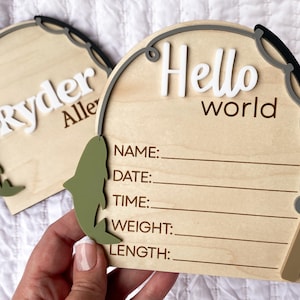 K&ouml;nnte beinhalten: Zwei Holzschilder zur Babyank&uuml;ndigung. Eines tr&auml;gt die Aufschrift "Ryder Allen" in wei&szlig;en Buchstaben. Das andere tr&auml;gt die Aufschrift "Hello world" in wei&szlig;en Buchstaben. Beide haben ein Angelruten-Design und Platz, um die Details des Babys zu notieren.