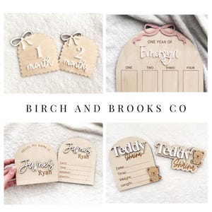 May include: A collection of light-colored wooden milestone and birth announcement plaques featuring arched shapes, white and pink 3D lettering, and engraved details. The plaques include text such as 1 month, 2 months, One Year of Emersyn, Hello My Name Is James Ryan, and Teddy Shaina with bear icons.