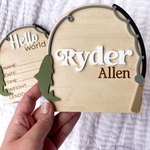 K&ouml;nnte beinhalten: Holzschild zur Babyank&uuml;ndigung mit dem Namen "Ryder Allen" in wei&szlig;en und braunen Buchstaben. Das Schild hat die Form einer Angelpose mit einem gr&uuml;nen Fischakzent und den Worten "Hello world" auf einem zweiten Schild.