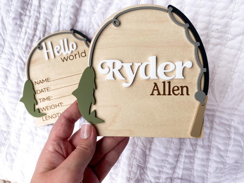 K&ouml;nnte beinhalten: Holzschild zur Babyank&uuml;ndigung mit dem Namen "Ryder Allen" in wei&szlig;en und braunen Buchstaben. Das Schild hat die Form einer Angelpose mit einem gr&uuml;nen Fischakzent und den Worten "Hello world" auf einem zweiten Schild.