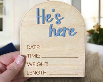 May include: A wooden sign with a blue inscription that reads "He's here". The sign has spaces to fill in the date, time, weight, and length of a newborn.