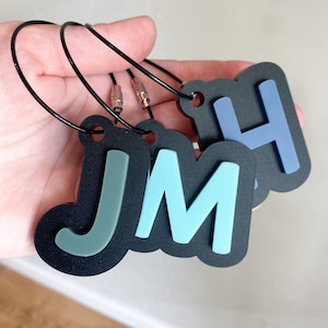 May include: Three black acrylic luggage tags featuring raised letters J, M, and H in muted sage green, light blue, and periwinkle blue. Each tag is attached to a thin, flexible black wire cable with a secure metal screw-top fastener for attachment.