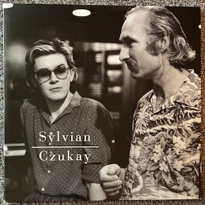 May include: Black and white album cover featuring two men. One man wears sunglasses and a polka dot shirt. The other man wears a patterned shirt. The text "Sylvian Czukay" is printed on the cover.