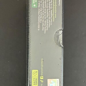 May include: A rectangular box for the Exploding Kittens card game. The box is dark with text that includes the game's name, age range, and player count. It also has a special code for a gift.